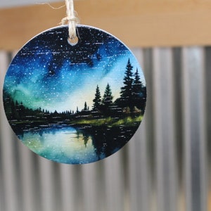 May include: A round wooden ornament with a painted scene of a forest lake at night. The sky is a deep blue with white stars and a green and blue aurora borealis. The lake is reflected in the water. The trees are silhouetted against the sky.