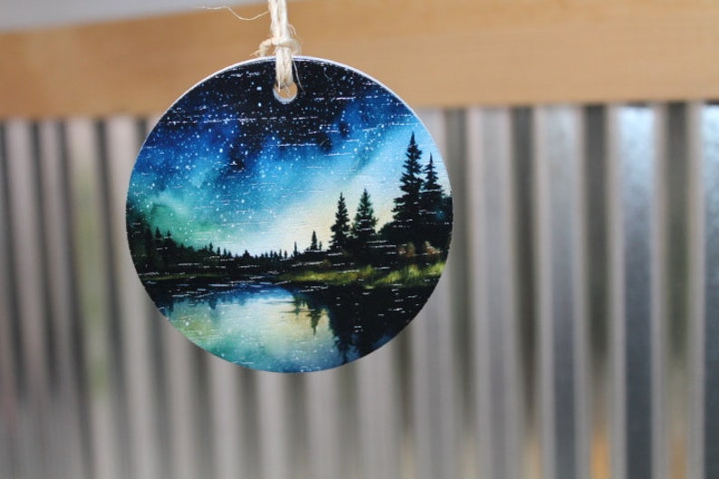 May include: A round wooden ornament with a painted scene of a forest lake at night. The sky is a deep blue with white stars and a green and blue aurora borealis. The lake is reflected in the water. The trees are silhouetted against the sky.