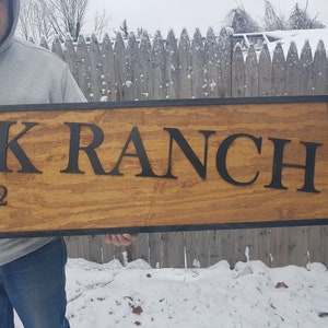 May include: A wooden sign with black lettering that reads "EK RANCH 1872". The sign has a black border and is held by a person.