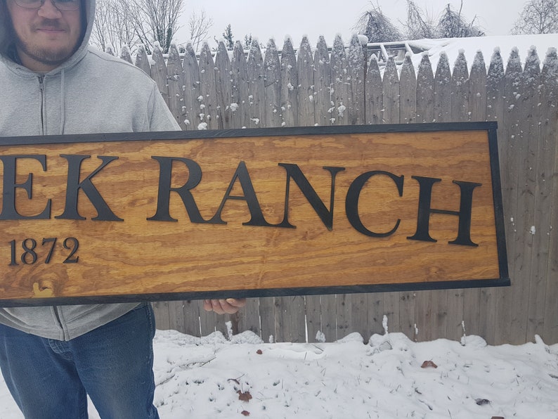 May include: A wooden sign with black lettering that reads "EK RANCH 1872". The sign has a black border and is held by a person.