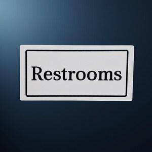 May include: A rectangular white sign with a black border and the word "Restrooms" in black, centered on the sign. The sign is against a dark blue background.