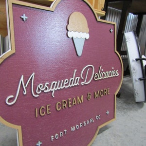 May include: A wooden sign with a maroon background and a gold border. The sign has a stylized white font that reads "Mosqueda Delicacies Ice Cream & More Fort Morgan, CO". There is a white and brown ice cream cone graphic in the upper left corner.
