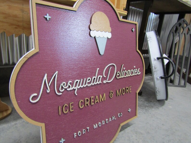 May include: A wooden sign with a maroon background and a gold border. The sign has a stylized white font that reads "Mosqueda Delicacies Ice Cream & More Fort Morgan, CO". There is a white and brown ice cream cone graphic in the upper left corner.