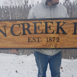 May include: A wooden sign with black lettering that reads "Twin Creek Ranch Est. 1872". The sign is held by a person in front of a snow-covered background.