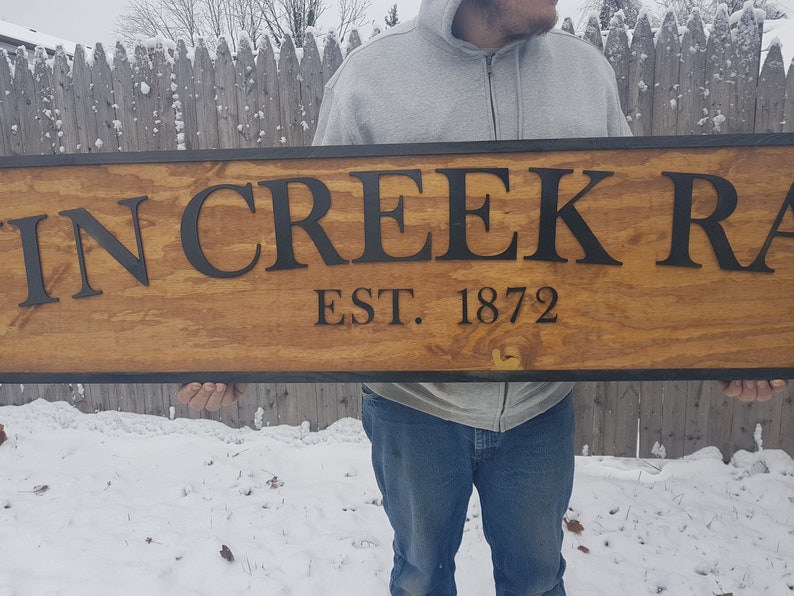 May include: A wooden sign with black lettering that reads "Twin Creek Ranch Est. 1872". The sign is held by a person in front of a snow-covered background.