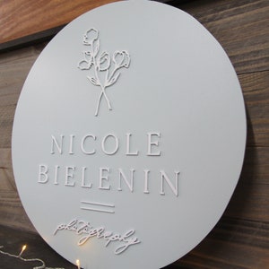 May include: A round, light blue sign with the name "Nicole Bielenin" in white, capital letters. The word "photography" is written in cursive below the name. A floral design is at the top of the sign.