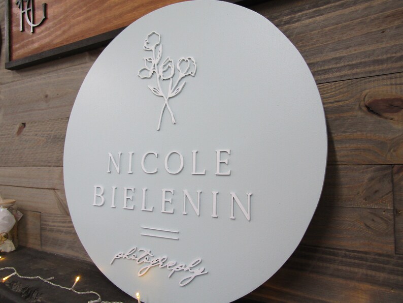 May include: A round, light blue sign with the name "Nicole Bielenin" in white, capital letters. The word "photography" is written in cursive below the name. A floral design is at the top of the sign.