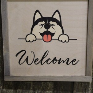 May include: A wooden sign with a gray frame features a cartoon black and white dog peeking over the top with its tongue sticking out. The sign says "Welcome" in black cursive lettering.