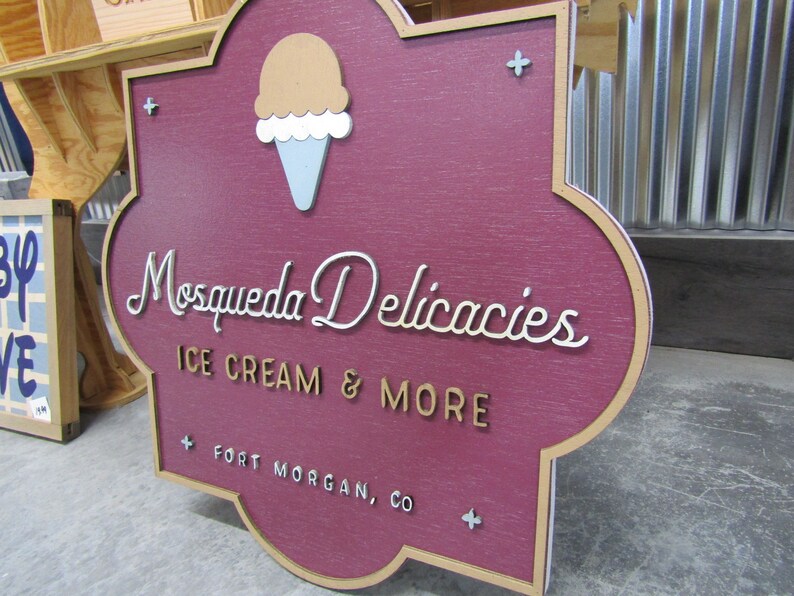 May include: A wooden sign with a maroon background and a gold border. The sign has a stylized ice cream cone graphic and the text "Mosqueda Delicacies Ice Cream & More Fort Morgan, CO".