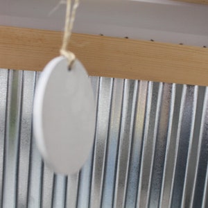 May include: A white ceramic ornament hangs from a string against a backdrop of corrugated metal and wood.