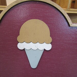 May include: A wooden sign with a red background and a white and brown ice cream cone cutout. The ice cream cone is in the center of the sign.