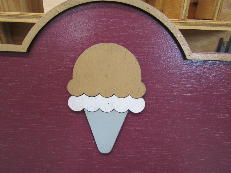 May include: A wooden sign with a red background and a white and brown ice cream cone cutout. The ice cream cone is in the center of the sign.