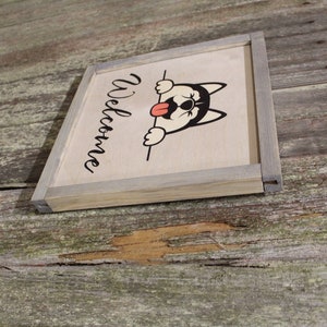 May include: A wooden sign with a gray frame and the word "Welcome" written in black cursive. A cartoon husky dog with a red tongue is peeking over the top of the sign.