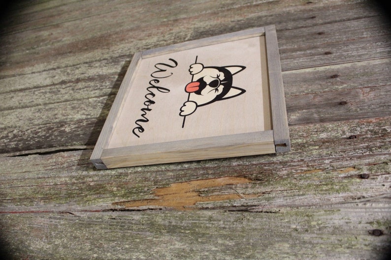 May include: A wooden sign with a gray frame and the word "Welcome" written in black cursive. A cartoon husky dog with a red tongue is peeking over the top of the sign.