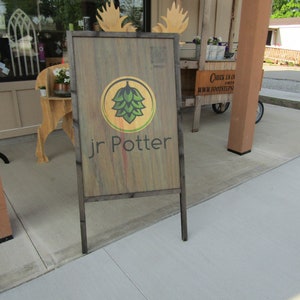 May include: A wooden sign with a black frame and a green hop leaf logo. The sign reads "Jr Potter" in black text.