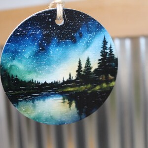 May include: Round wooden ornament with a painted scene of a lake, forest, and starry night sky. The sky is a gradient of blue, green, and purple with white stars. The lake is a dark blue with a reflection of the sky. The forest is a silhouette of dark green trees.