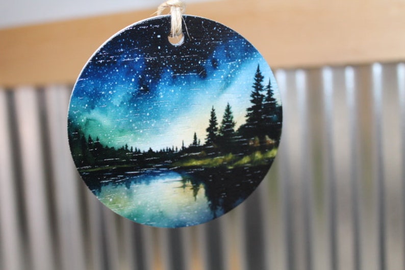 May include: Round wooden ornament with a painted scene of a lake, forest, and starry night sky. The sky is a gradient of blue, green, and purple with white stars. The lake is a dark blue with a reflection of the sky. The forest is a silhouette of dark green trees.