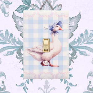 May include: A decorative light switch cover featuring a watercolor illustration of a goose wearing a blue bonnet. The cover has a blue and white gingham pattern and a gold-colored switch. Pink lace trim borders the top and bottom.