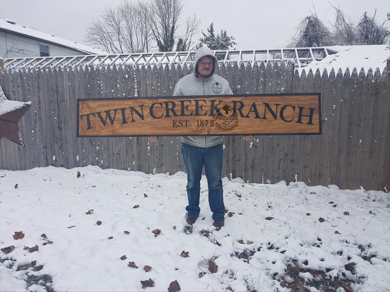 May include: A wooden sign with the text "Twin Creek Ranch Est. 1877" in black lettering. The sign is held by a person in front of a wooden fence and snow-covered ground.