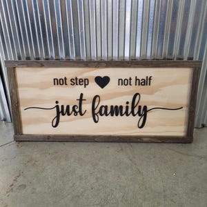 May include: Wooden sign with a light wood background and a dark brown frame. The sign features the words "not step", a black heart, "not half", and "just family" in a black script font. The sign is rectangular.