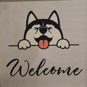 May include: A wooden sign with a black and white cartoon dog peeking over the top. The dog has a pink tongue sticking out and the sign says "Welcome".
