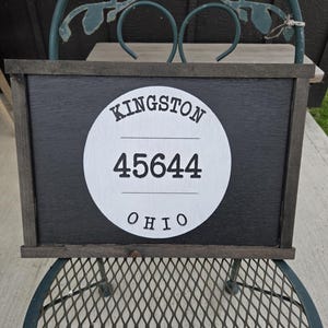 May include: A rectangular wooden sign with a dark gray background and a white circle in the center. The circle contains the text "KINGSTON", "45644", and "OHIO". The sign is framed with dark wood and sits on a metal chair.