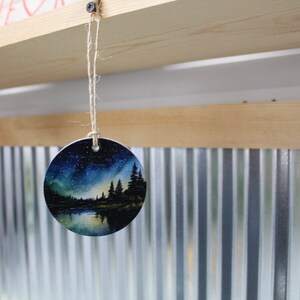 May include: A round, white ceramic ornament with a painted scene of a lake, trees, and a starry night sky. The ornament is hanging from a string.
