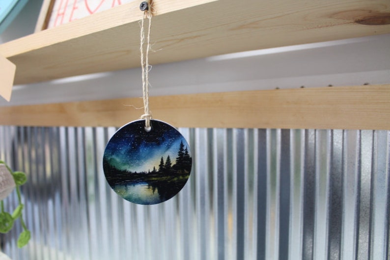 May include: A round, white ceramic ornament with a painted scene of a lake, trees, and a starry night sky. The ornament is hanging from a string.
