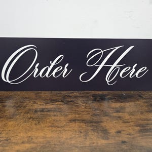 May include: A rectangular, navy blue sign with the words "Order Here" in elegant white script. The sign is angled and sits on a wooden surface, suitable for a business or event.