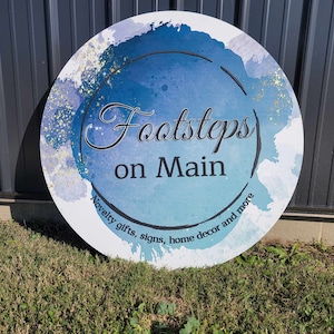 May include: A round sign with the text "Footsteps on Main" in a stylized font, set against a blue and white watercolor background with gold accents. The sign also includes the text "Novelty gifts, signs, home decor and more".
