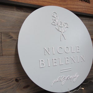 May include: A round, light blue sign with the name "NICOLE BIELENIN" in raised white lettering. Below the name is the word "photography" in a cursive font. A floral design is at the top.