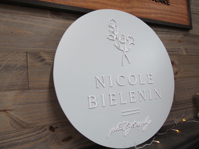 May include: A round, light blue sign with the name "NICOLE BIELENIN" in raised white lettering. Below the name is the word "photography" in a cursive font. A floral design is at the top.