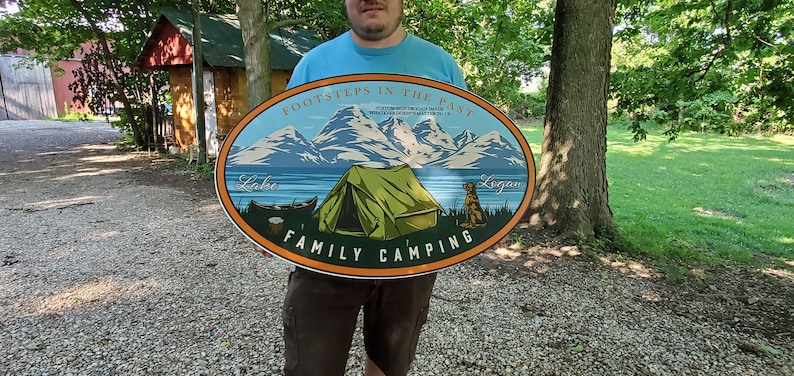 May include: A wooden oval sign with a blue and green mountain scene, a green tent, and a dog. The sign reads "Footsteps in the Past", "Lake", "Logan", and "Family Camping".