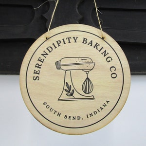 May include: A round wooden sign with the text "Serendipity Baking Co. South Bend, Indiana" and a black and white illustration of a stand mixer.