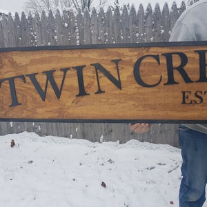 May include: A wooden sign with black lettering that reads "Twin Creek Est. 19..." The sign is held by a person's hand and has a black frame.