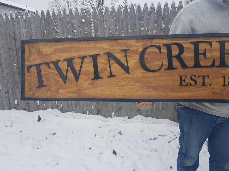 May include: A wooden sign with black lettering that reads "Twin Creek Est. 19..." The sign is held by a person's hand and has a black frame.