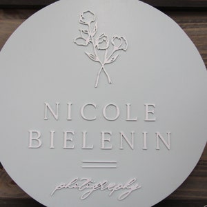 May include: A round, light blue sign with the name "NICOLE BIELENIN" in capital letters. Below the name are two horizontal lines, and the word "photography" is written in cursive. A floral design is at the top.