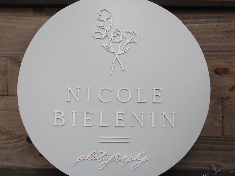 May include: A round, light blue sign with the name "NICOLE BIELENIN" in capital letters. Below the name are two horizontal lines, and the word "photography" is written in cursive. A floral design is at the top.