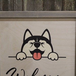 May include: A wooden sign with a gray frame features a cartoon husky dog with a black and white face and a pink tongue sticking out. The dog is peeking over a sign that says "Welcome".