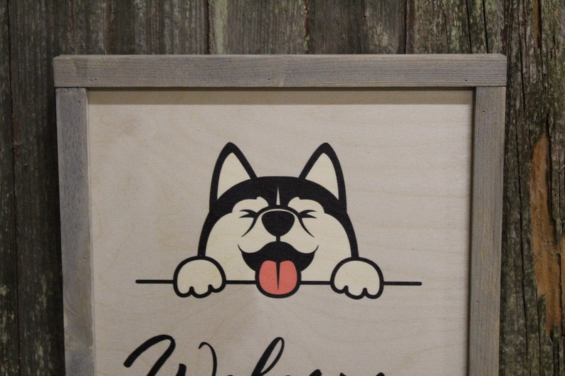 May include: A wooden sign with a gray frame features a cartoon husky dog with a black and white face and a pink tongue sticking out. The dog is peeking over a sign that says "Welcome".
