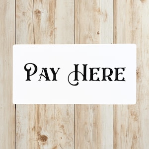 May include: A white rectangular sign with the words "PAY HERE" in black, elegant, serif font. The sign is set against a light brown, wooden background with vertical planks.