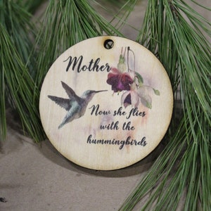 May include: A round wooden ornament with a hummingbird and flowers painted on it. The ornament has the text "Mother Now she flies with the hummingbirds" printed on it.