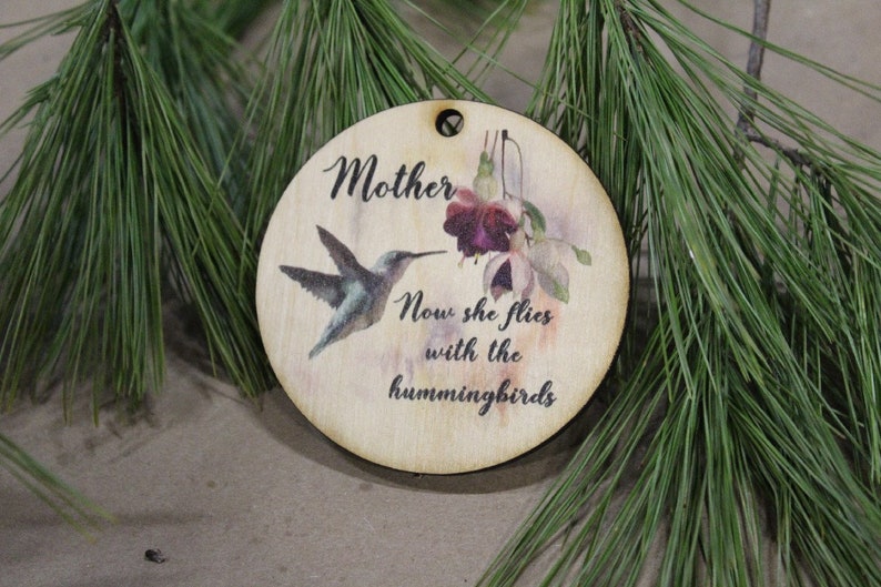 May include: A round wooden ornament with a hummingbird and flowers painted on it. The ornament has the text "Mother Now she flies with the hummingbirds" printed on it.