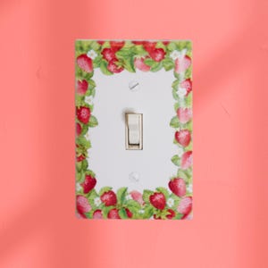 May include: A white light switch cover with a decorative border of red strawberries, green leaves, and white flowers. The switch itself is a light beige color, and the cover is mounted on a coral-colored wall.