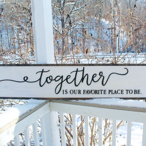 May include: White wooden sign with black cursive lettering that reads "together is our favorite place to be." The sign is leaning against a white railing with a snowy background.
