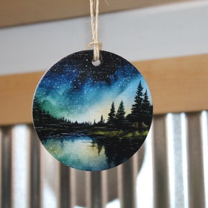 May include: A round wooden ornament with a painted scene of a forest at night with a starry sky and a lake reflecting the stars. The ornament is painted in shades of blue, green, and black.