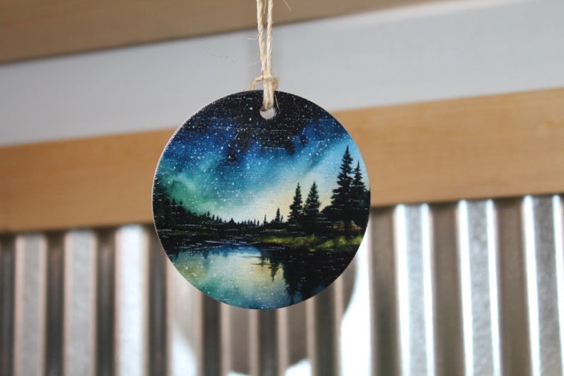 May include: A round wooden ornament with a painted scene of a forest at night with a starry sky and a lake reflecting the stars. The ornament is painted in shades of blue, green, and black.