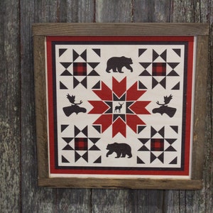 May include: A framed wall art piece with a red, black, and white quilt pattern featuring a red starburst in the center. The quilt pattern includes silhouettes of bears, moose, and deer.