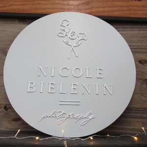 May include: A round, light gray sign with the name "NICOLE BIELENIN" in raised white lettering. Below the name is the word "photography" in cursive. The top of the sign features a floral design. The sign is mounted on a wooden background.