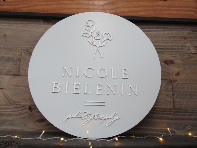 May include: A round, light gray sign with the name "NICOLE BIELENIN" in raised white lettering. Below the name is the word "photography" in cursive. The top of the sign features a floral design. The sign is mounted on a wooden background.